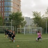 Grupă de copii care fac exerciții cu mingea pe un teren de sport, sub îndrumarea antrenorului