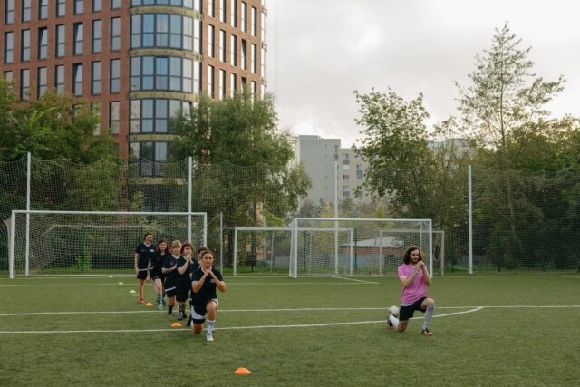 Grupă de copii care fac exerciții cu mingea pe un teren de sport, sub îndrumarea antrenorului