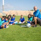 Antrenor care notează planul de pregătire pe un clipboard, lângă pista de atletism a unui stadion